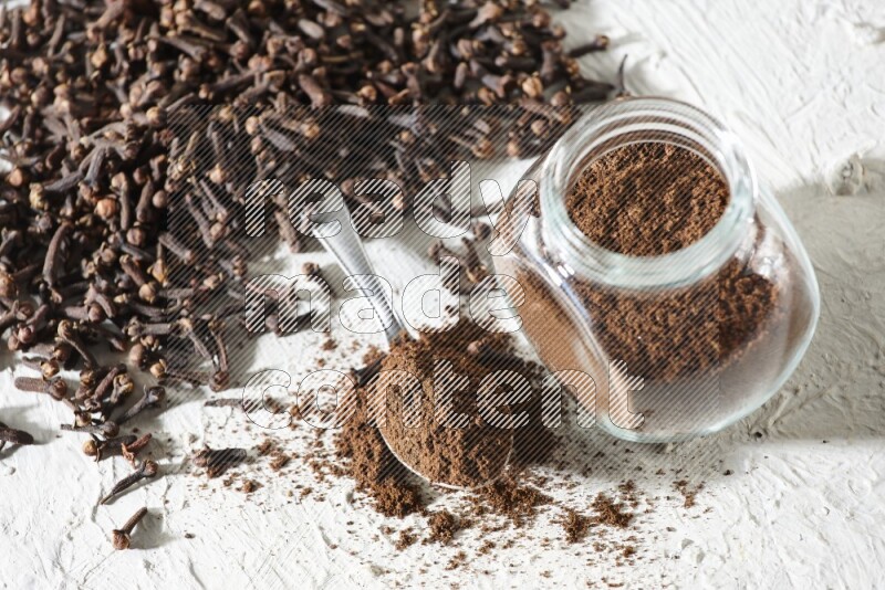A glass spice jar and a metal spoon full of cloves powder and cloves spread on textured white flooring