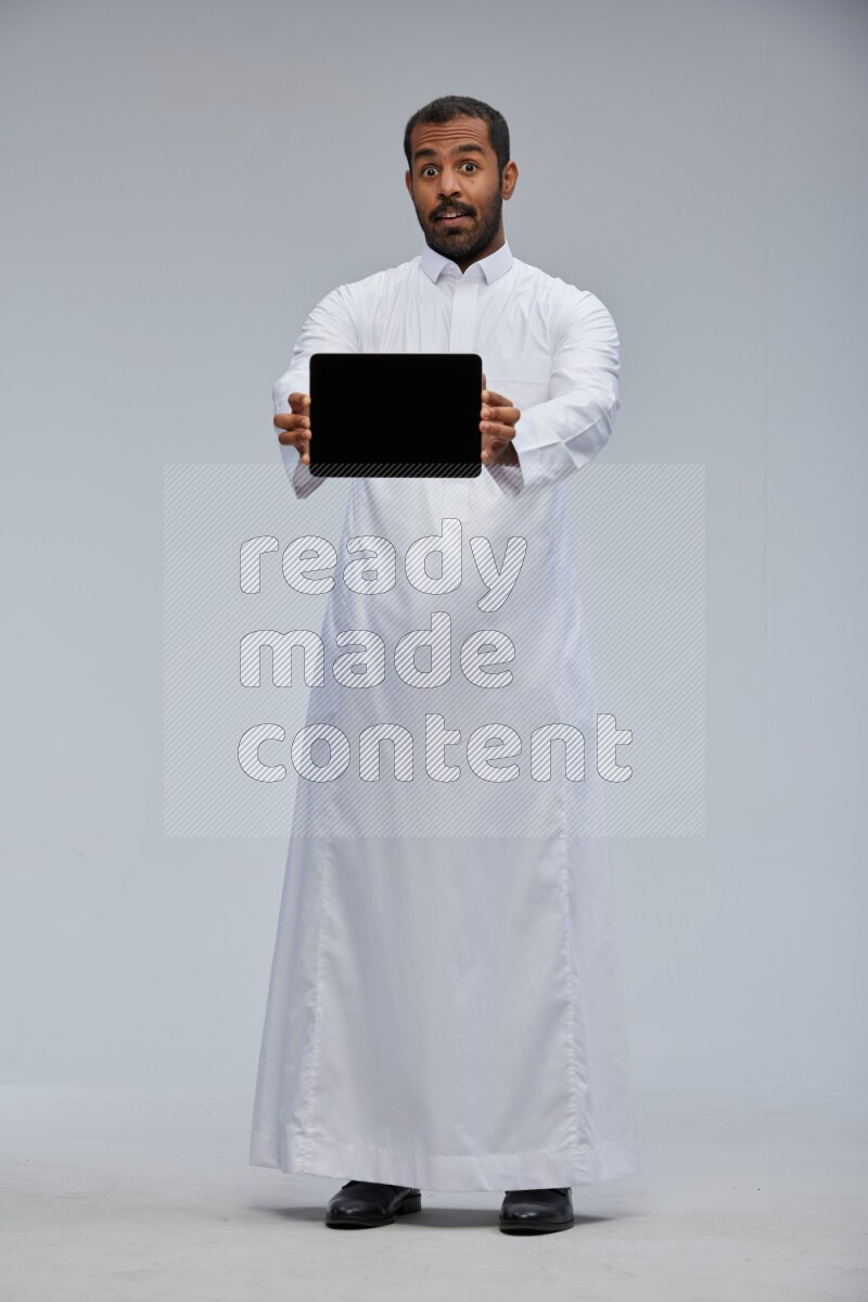 Saudi man wearing Thob standing showing tablet to camera on Gray background