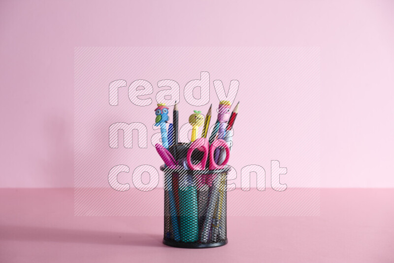 Pens, pencils and other different school supplies in a metal pen holder on rose background