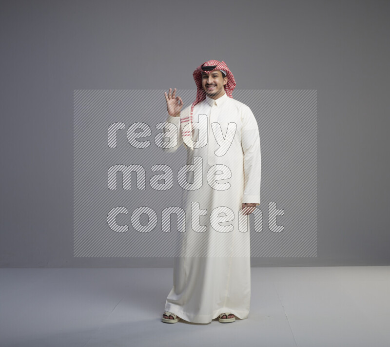 A Saudi man standing wearing thob and red shomag interacting with the camera on gray background