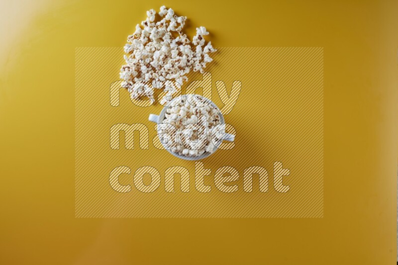 A white ceramic bowl full of popcorn with popcorn beside it on a yellow background in a top view shot