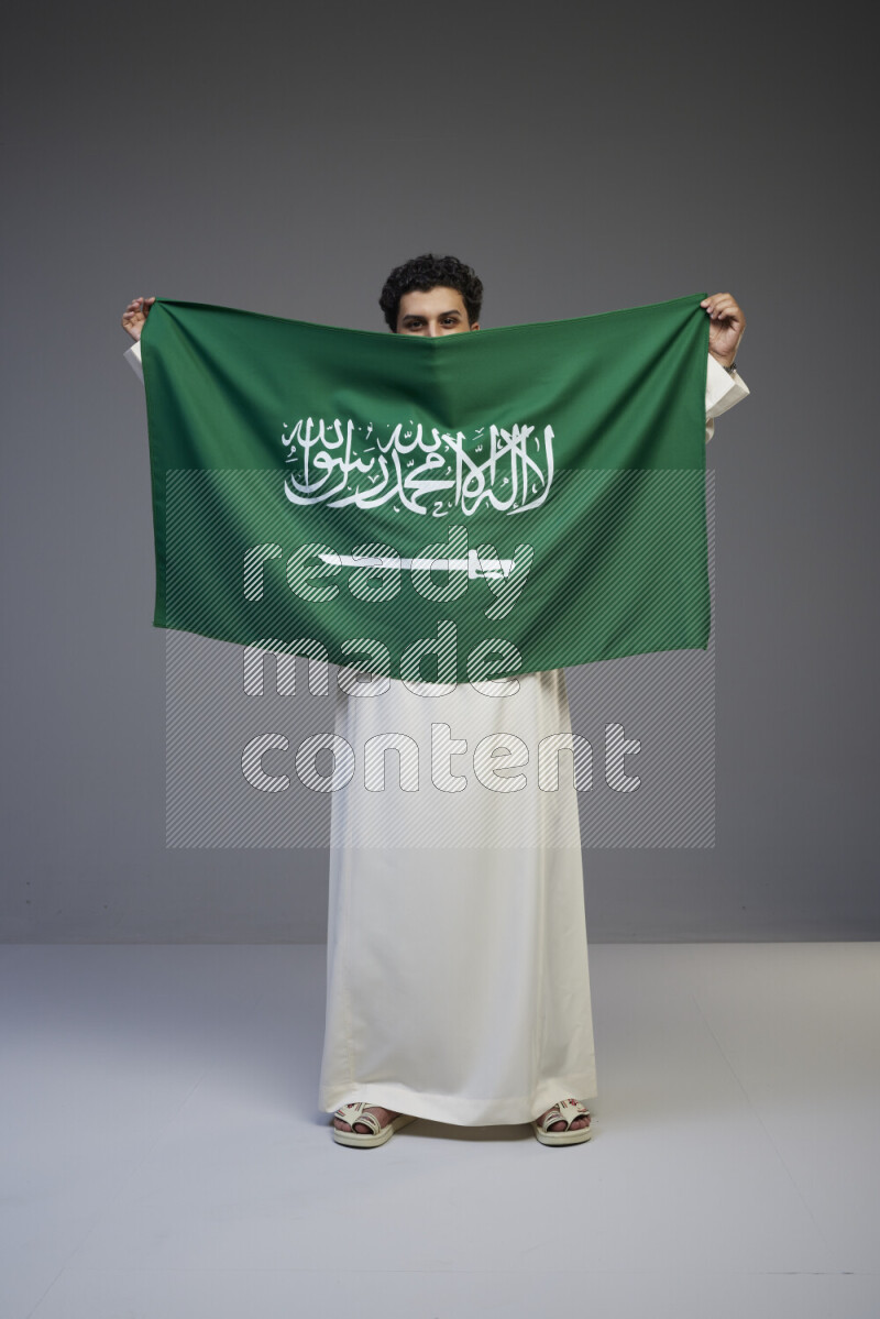 A Saudi man standing wearing thob holding big Saudi flag on gray background
