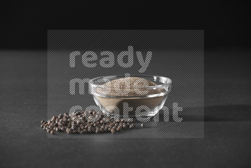 A glass bowl full of powder black pepper powder and black pepper beads on the floor on black flooring