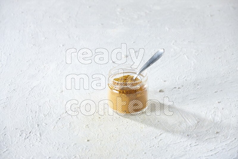 A glass jar and a metal spoon full of turmeric powder on a textured white flooring