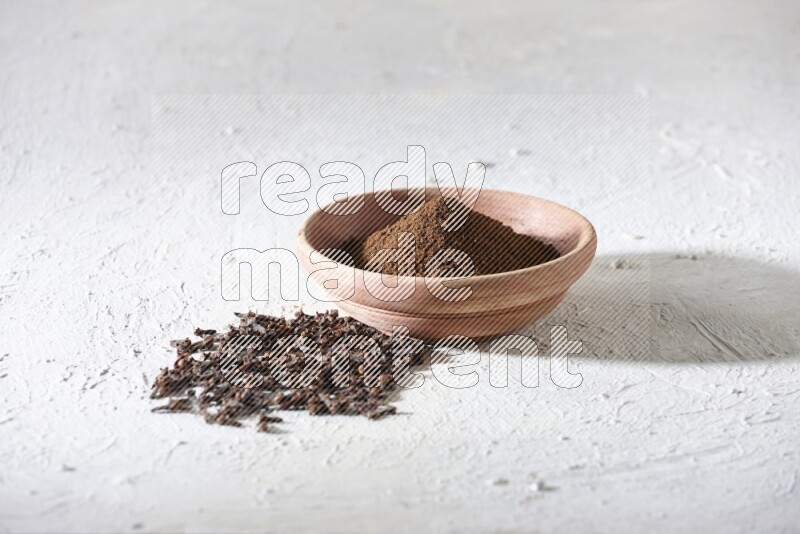A wooden bowl full of cloves powder with whole cloves beside it on a textured white flooring