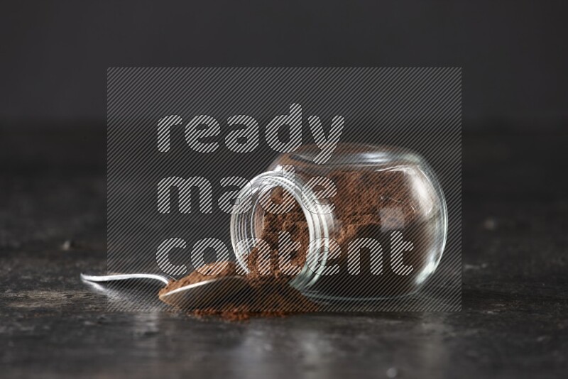 A flipped glass spice jar and a metal spoon full of cloves powder on textured black flooring