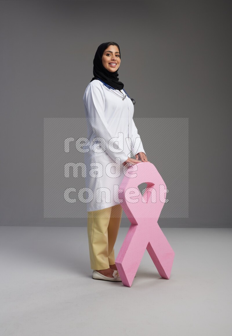 Saudi woman wearing lab coat with stethoscope standing holding awareness ribbon symbol standing on Gray background
