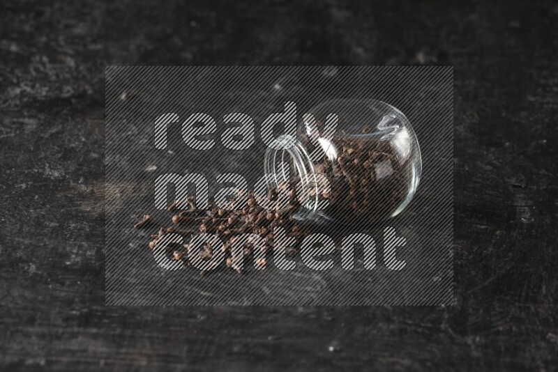 A flipped glass spice jar full of cloves and cloves came out of it on textured black flooring