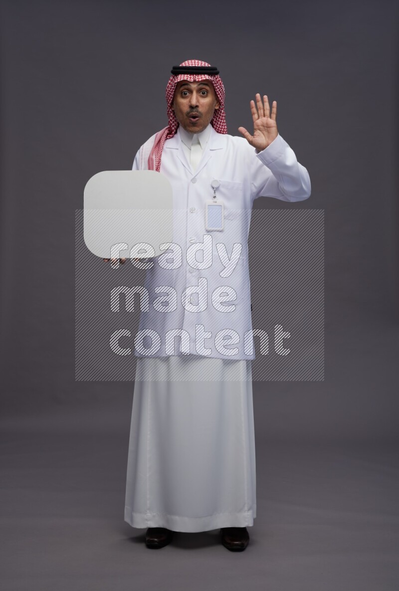 Saudi man wearing thob with lab coat and shomag with pocket employee badge standing holding social media sign on gray background