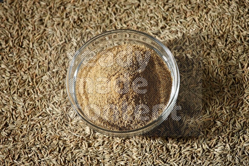A glass bowl full of cumin powder surrounded by cumin seeds on black flooring