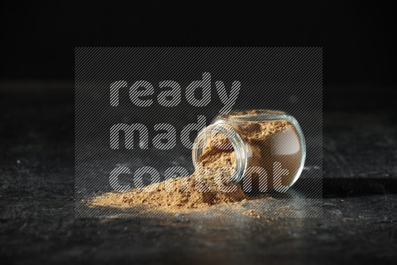 A flipped glass spice jar full of allspice powder and powder spilled out of it on a textured black flooring