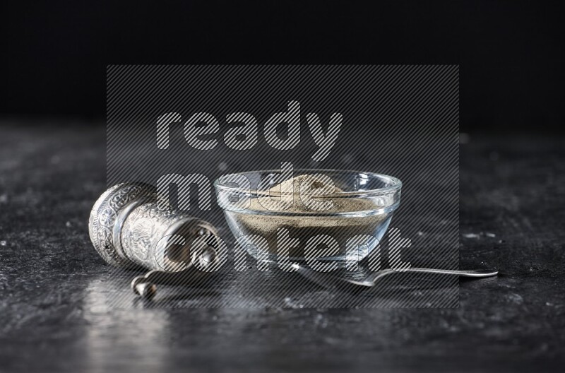 A glass bowl full of white pepper powder with pepper beads, a metal grinder and a metal spoon on textured black flooring