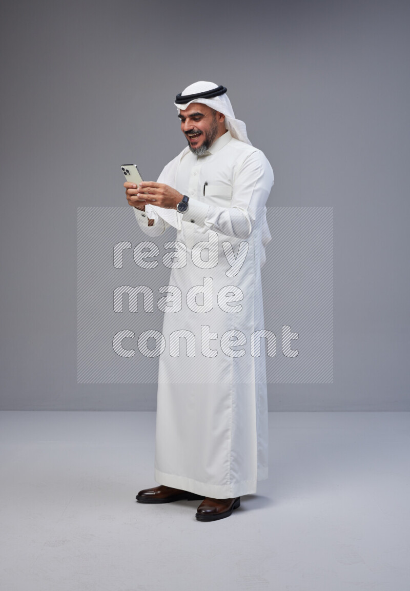 Saudi man Wearing Thob and white Shomag standing texting on phone on Gray background