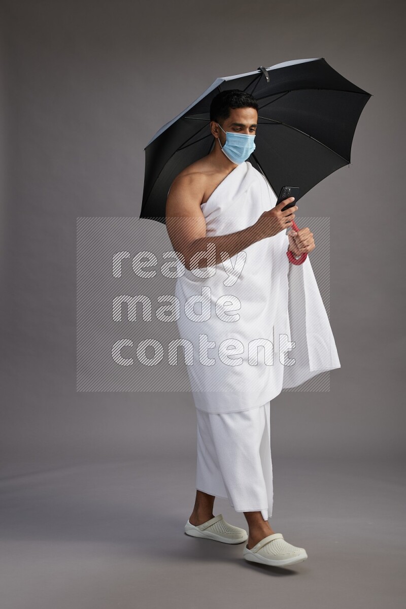 A man wearing Ehram and mask Standing holding umbrella on gray background on gray background
