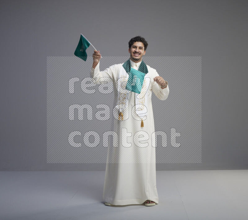 A Saudi man standing wearing thob and saudi flag scarf and holding small saudi flag on gray background