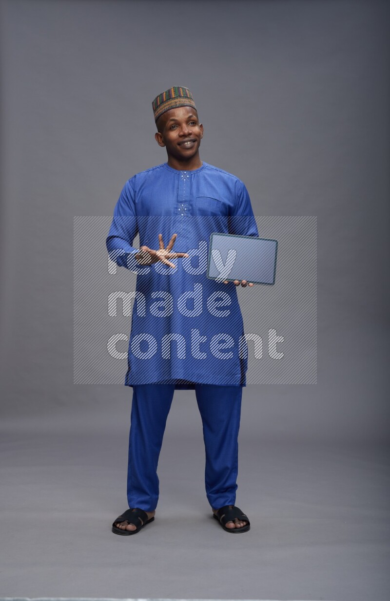 Man wearing Nigerian outfit standing showing tablet to camera on gray background