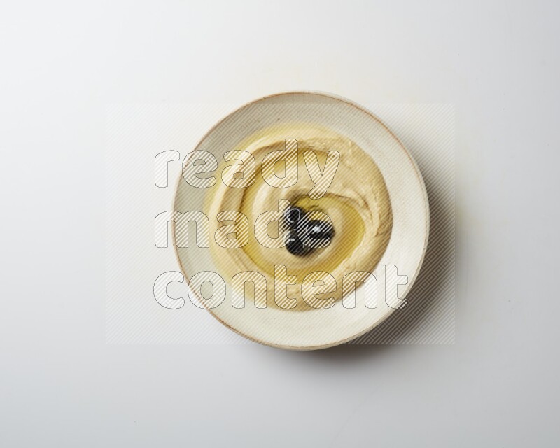 Hummus in a pottry plate garnished with black olives on a white background