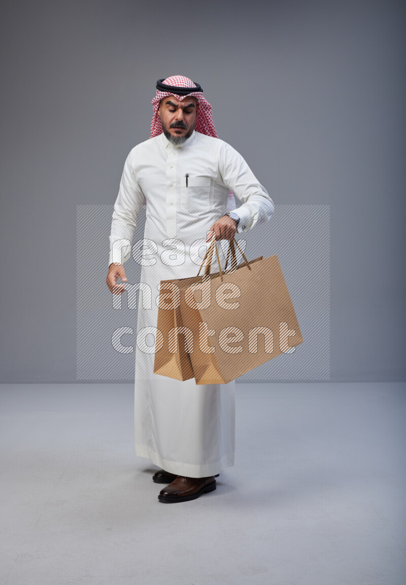 Saudi man Wearing Thob and red Shomag standing holding shopping bag on Gray background
