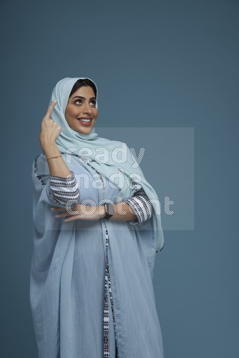 A Saudi woman pointing in a blue background wearing a blue Abaya with hijab