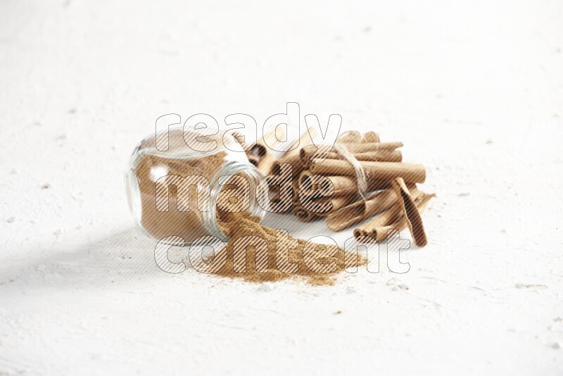 Flipped herbs glass jar full of cinnamon powder and cinnamon sticks in the back on a textured white background