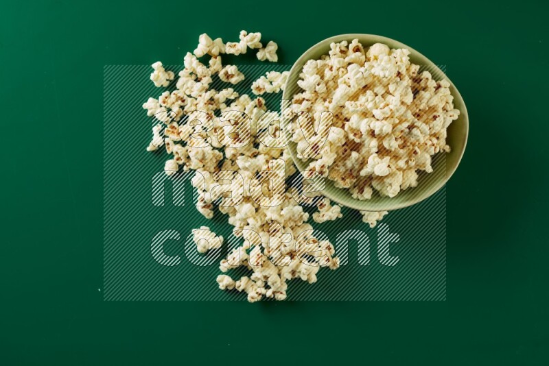 A green ceramic bowl full of popcorn with popcorn beside it on a green background in a top view shot