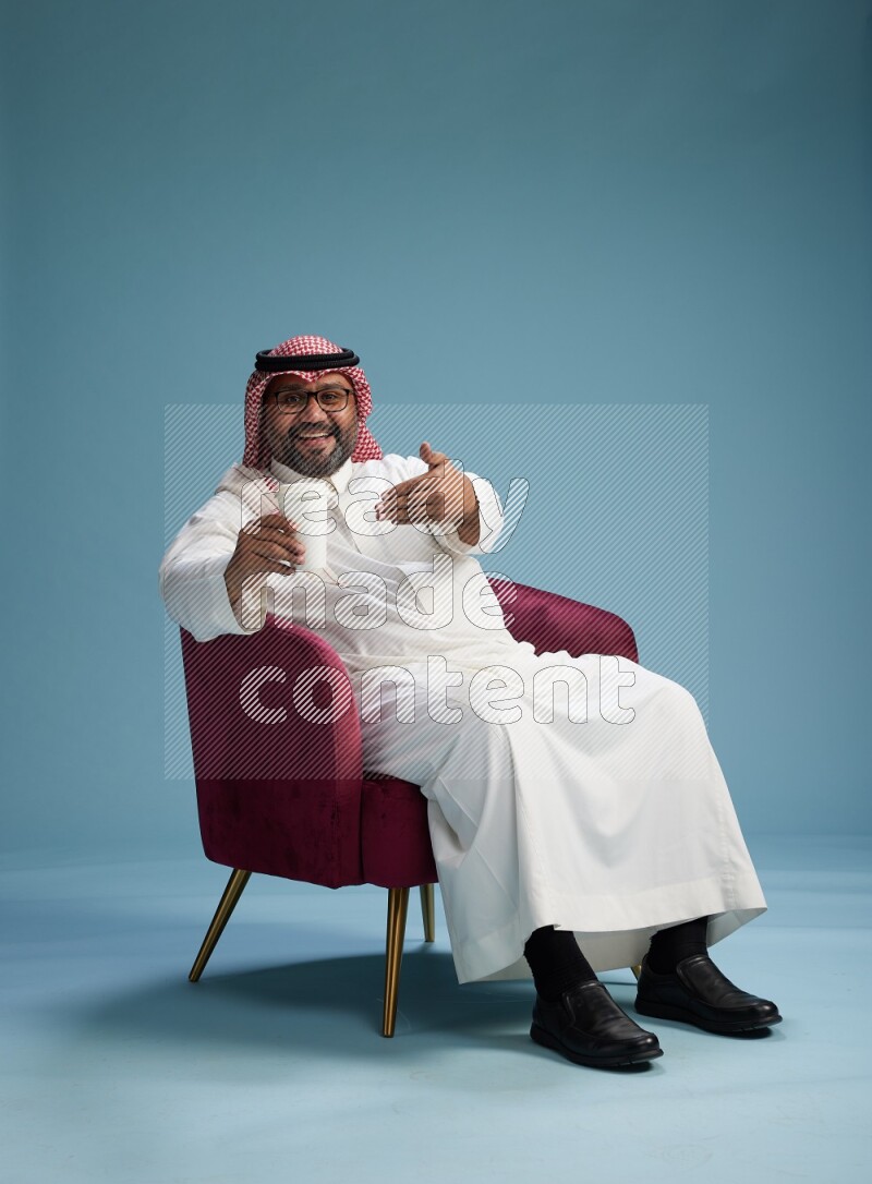 Saudi Man with shimag sitting on chair drinking coffee on blue background