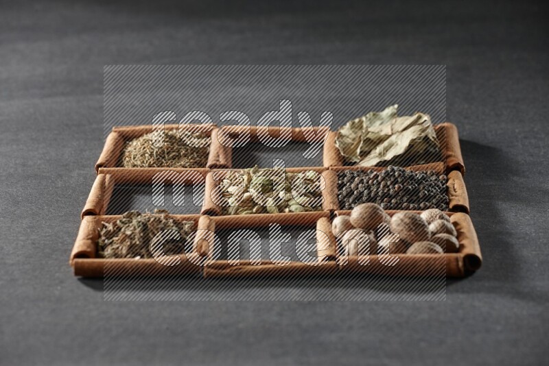 9 مربعات من أعواد القرفة ممتلئة بالهيل في المنتصف محاطة بجوزة الطيب والفلفل الأسود وورق الغار والبهارات والكمون والفلفل الأبيض والريحان المجفف والملح على أرضية سوداء