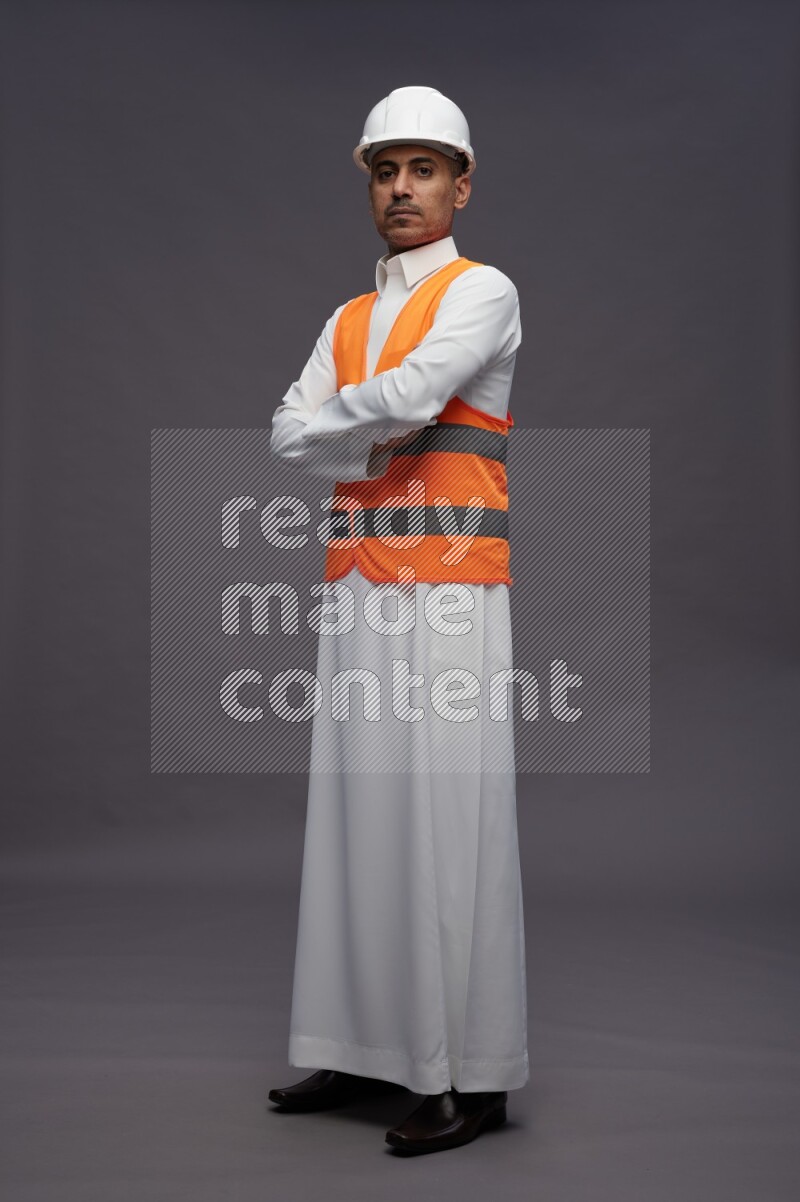 Saudi man wearing thob with engineer vest standing with crossed arms on gray background