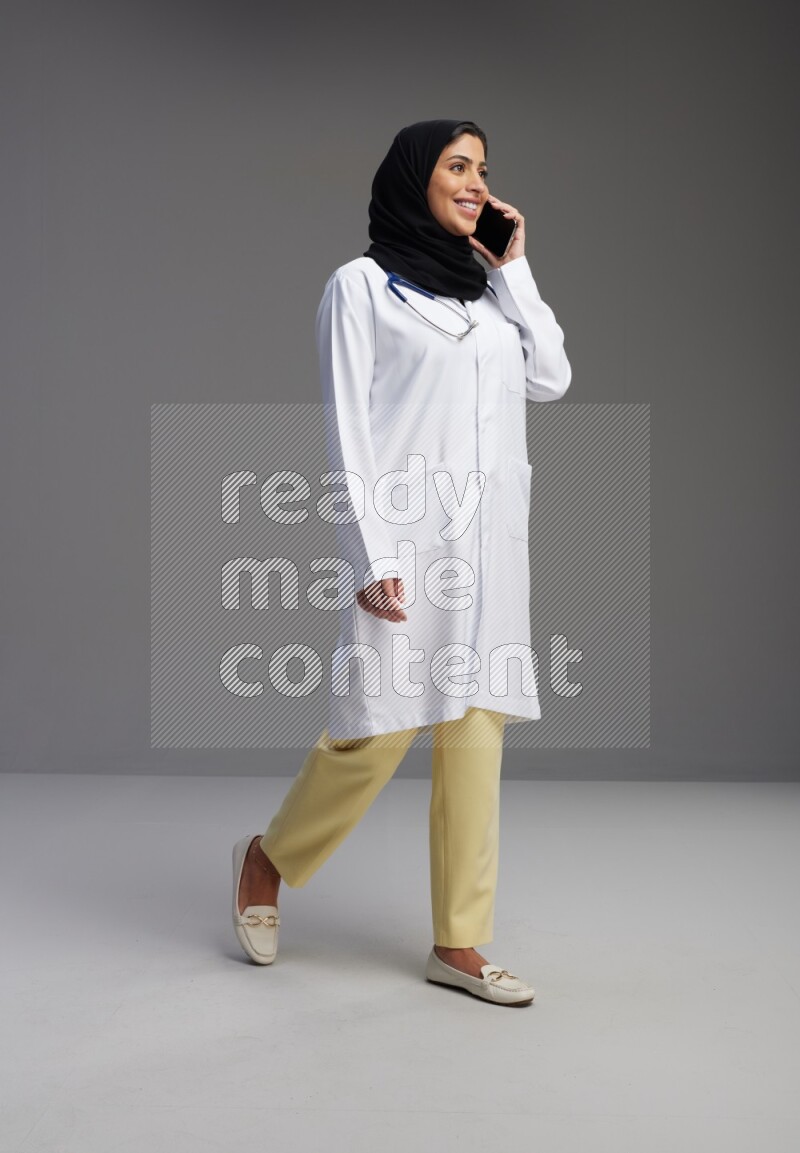Saudi woman wearing lab coat with stethoscope standing talking on phone on Gray background