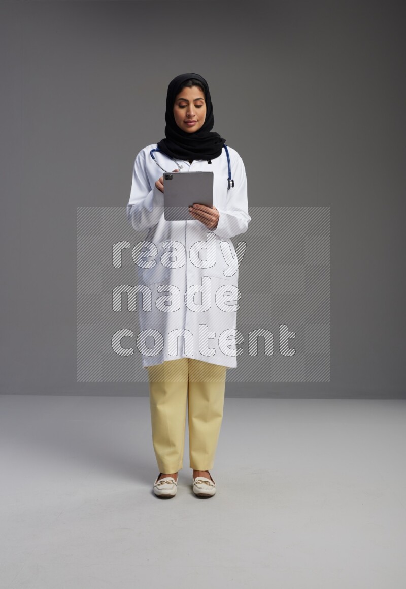 Saudi woman wearing lab coat with stethoscope standing working on tablet on Gray background