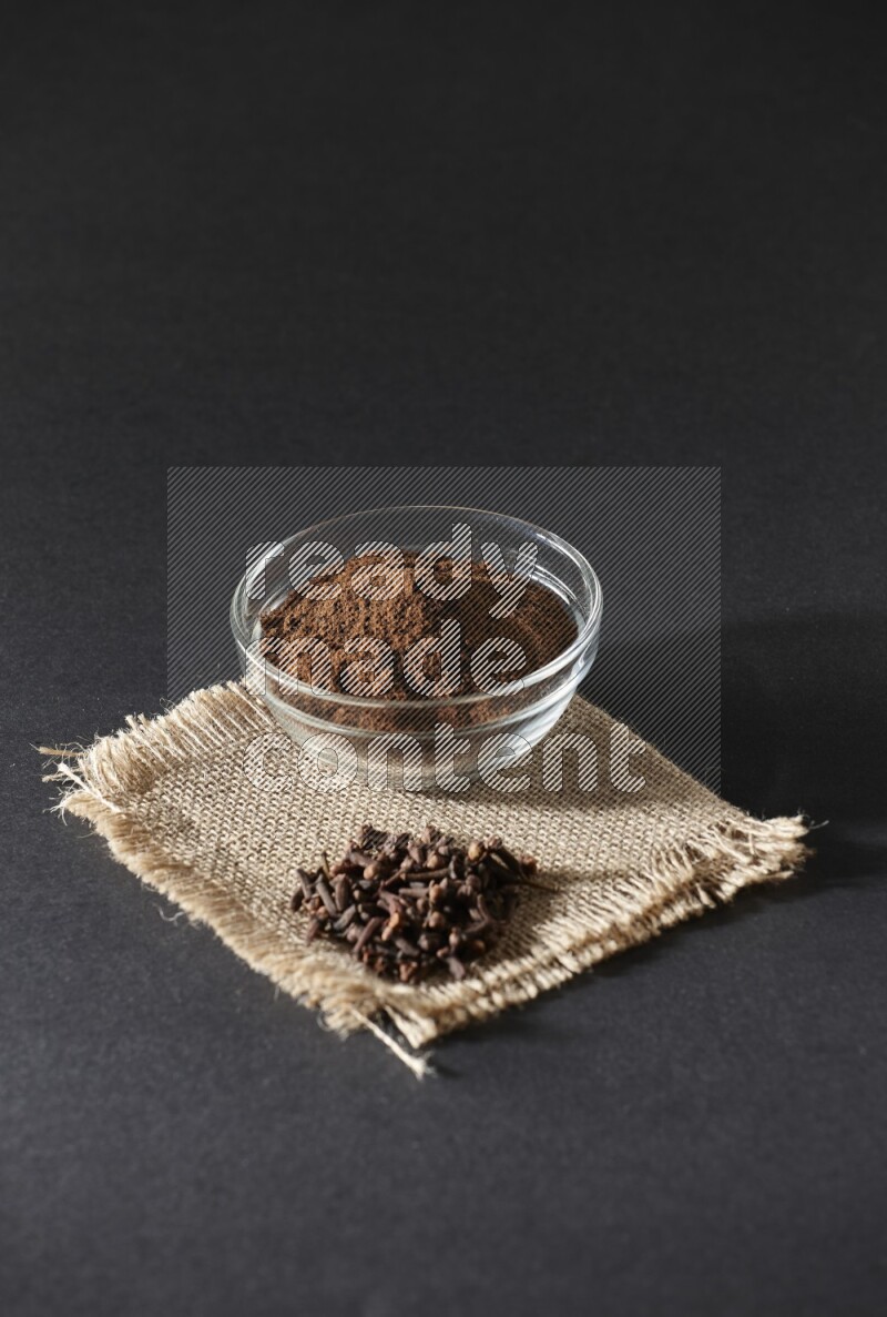 A glass bowl full of cloves powder with cloves grains on a burlap piece on a black flooring