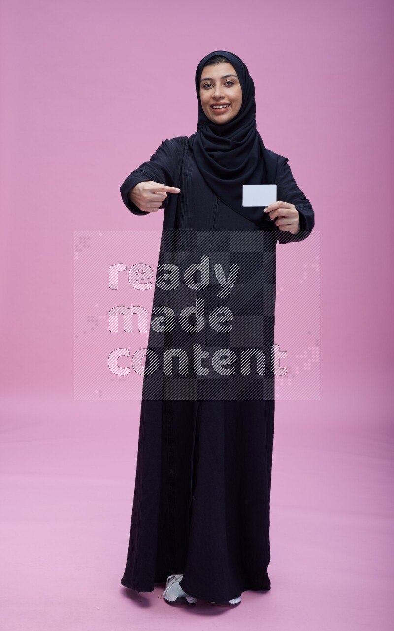 Saudi woman wearing Abaya standing holding ATM card on pink background
