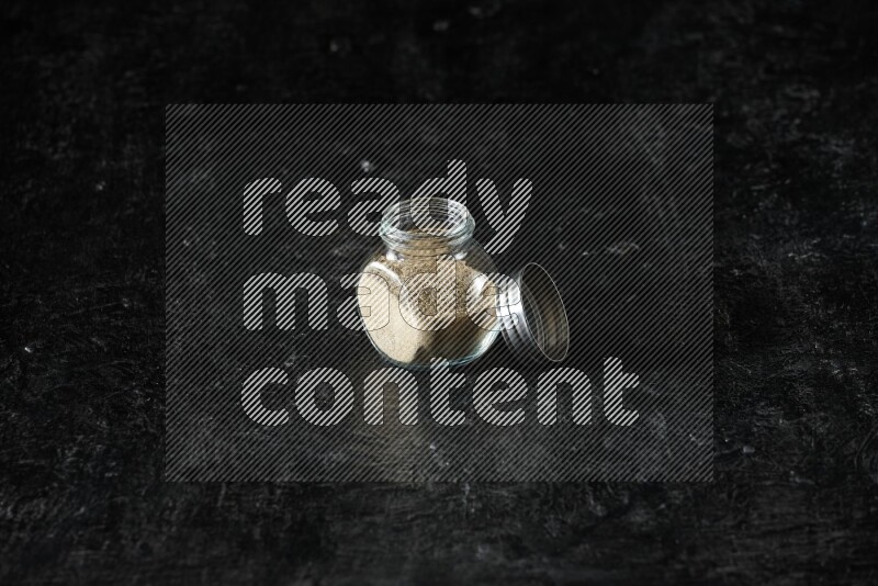 A glass spice jar full of cardamom powder on textured black flooring
