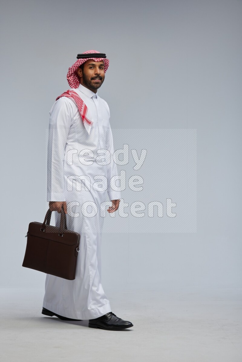 Saudi man Wearing Thob and shomag standing holding bag on Gray background