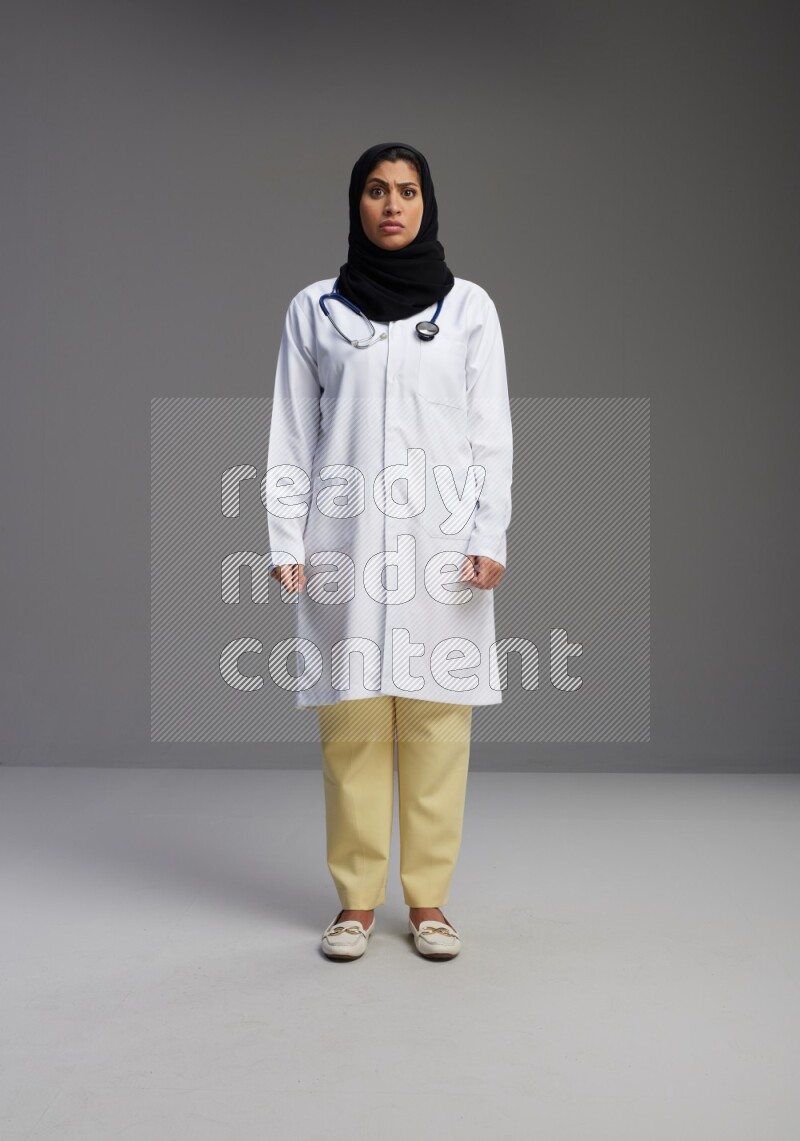 Saudi woman wearing lab coat with stethoscope standing interacting with the camera on Gray background