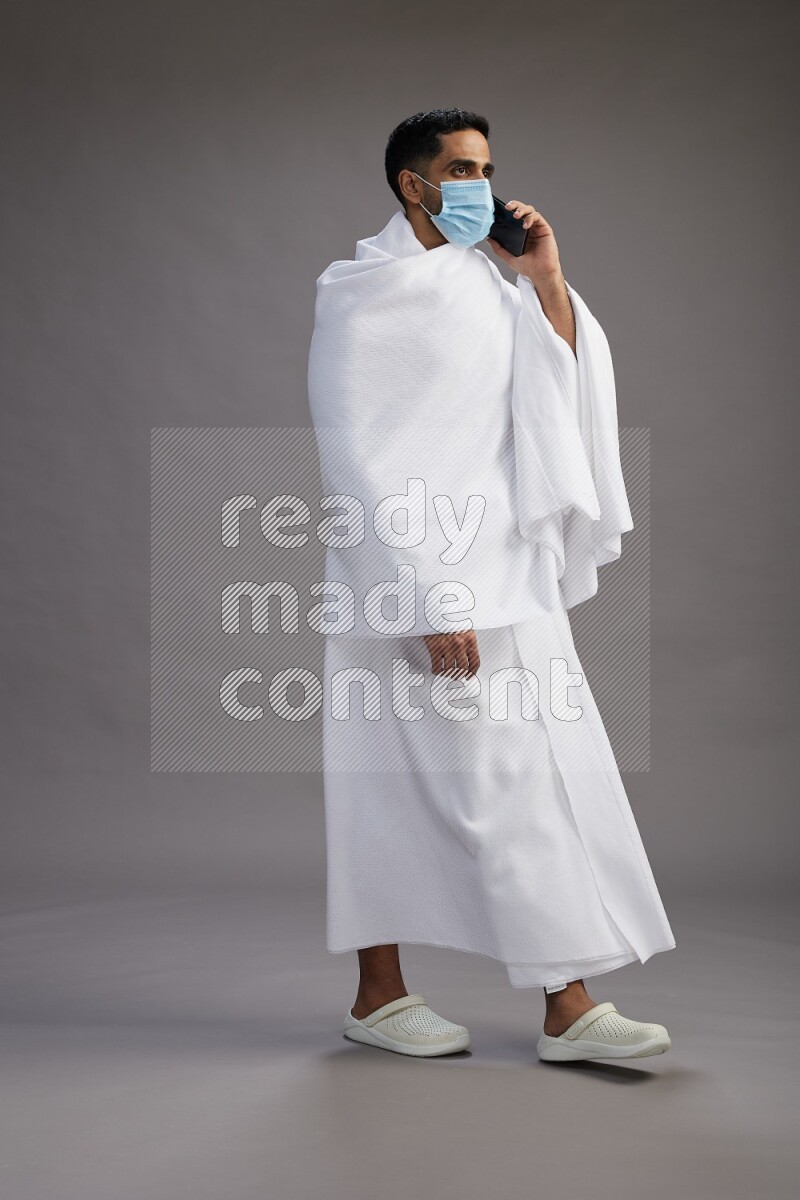 A man wearing Ehram with face mask Standing talking on phone on gray background