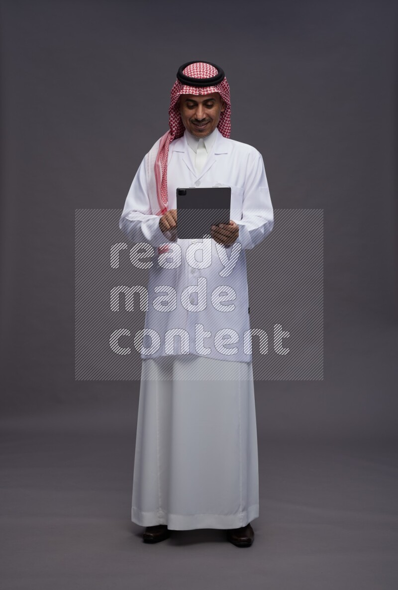 Saudi man wearing thob with lab coat and shomag with pocket employee badge standing working on tablet on gray background