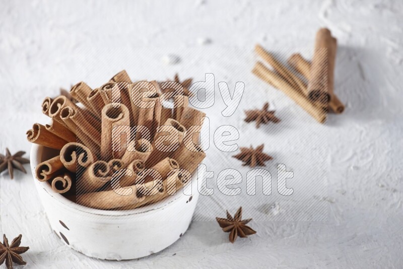 White bowl full of cinnamon sticks surrounded by star anis on a textured white background in different angles