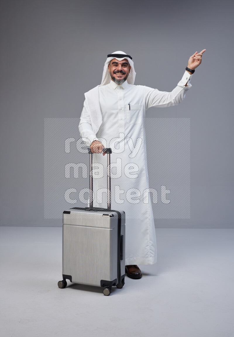 Saudi man wearing Thob and white Shomag standing holding Travel bag on Gray background