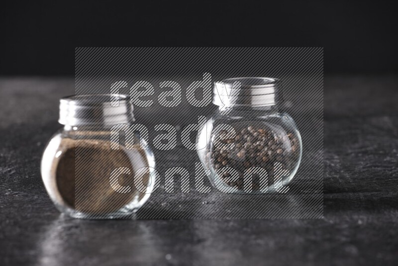 2 glass spice jars full of black pepper powder and black pepper beads on textured black flooring