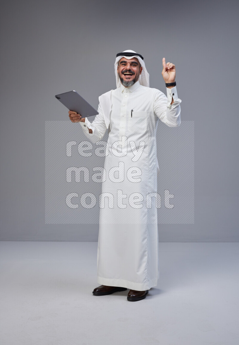 Saudi man Wearing Thob and white Shomag standing working on tablet on Gray background