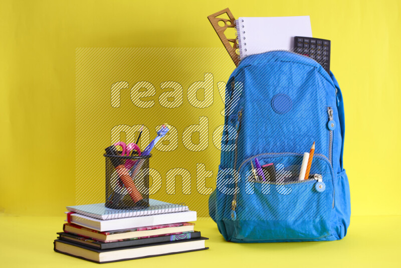 A school bag with assorted school supplies in and beside it on yellow background