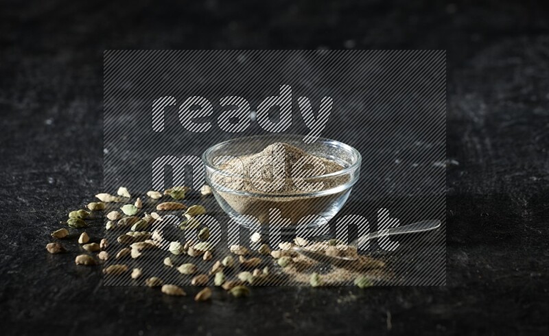 A glass bowl and a metal spoon full of cardamom powder with cardamom seeds beside them on textured black flooring