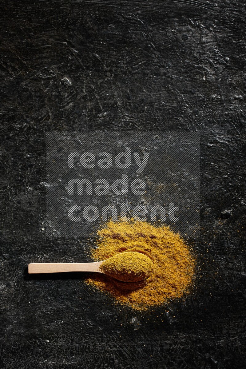 A wooden spoon full of turmeric powder on textured black background