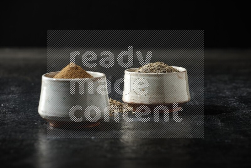 2 beige bowls, one full of cumin powder and the other full of seeds on a textured black flooring