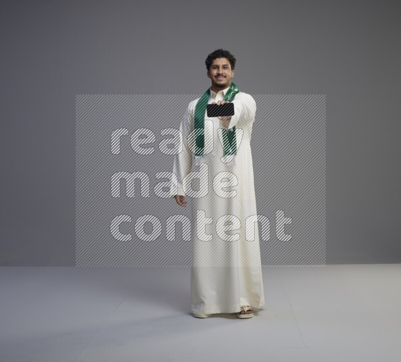 A Saudi man standing wearing thob and Saudi flag scarf showing phone to camera on gray background