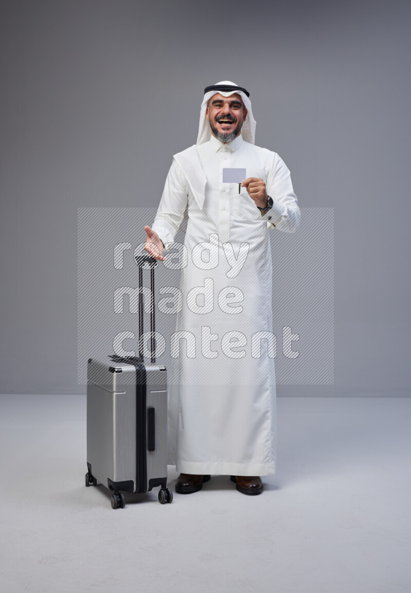 Saudi man wearing Thob and white Shomag standing holding Travel bag and ATM card on Gray background