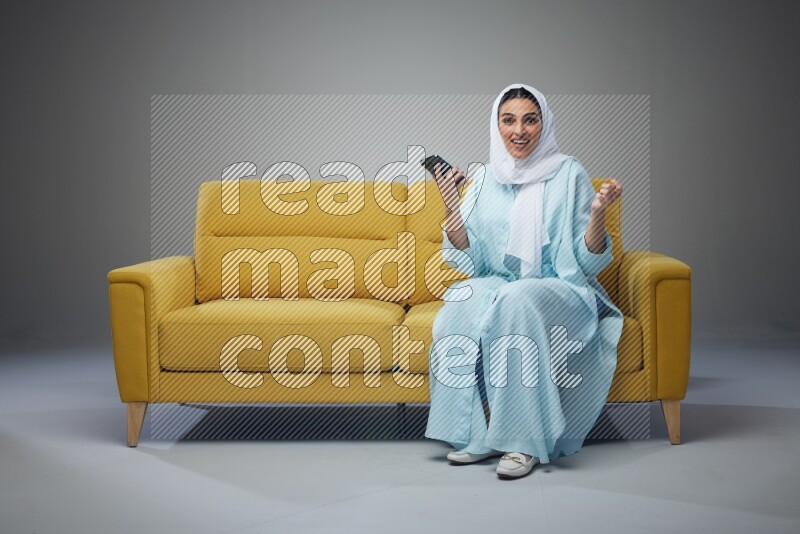 A Saudi woman wearing a light blue Abaya and white head scarf sitting on a yellow sofa and texting on her phone on a grey background