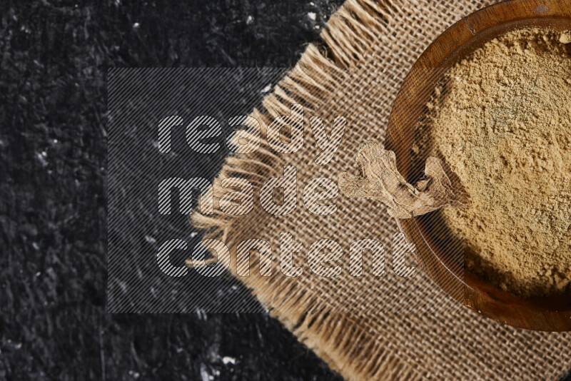 A wooden bowl full of ground ginger powder with a wooden spoon on it all on a burlap fabric on black background