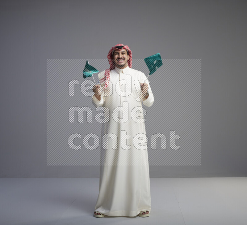 A Saudi man standing wearing thob and red shomag raising small Saudi flag on gray background